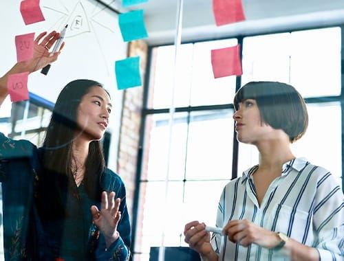 two women colleagues brainstorming meeting office large windows glass post it notes