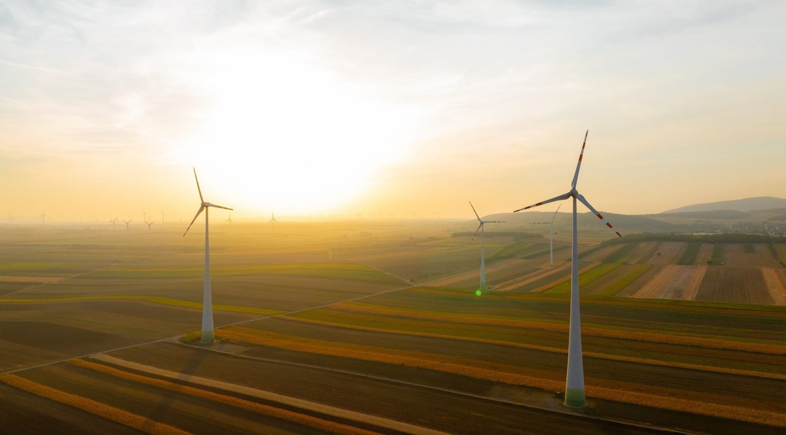 Wind turbines in ploughed fields. The sun is beginning to set on the horizon in the top left of the image.